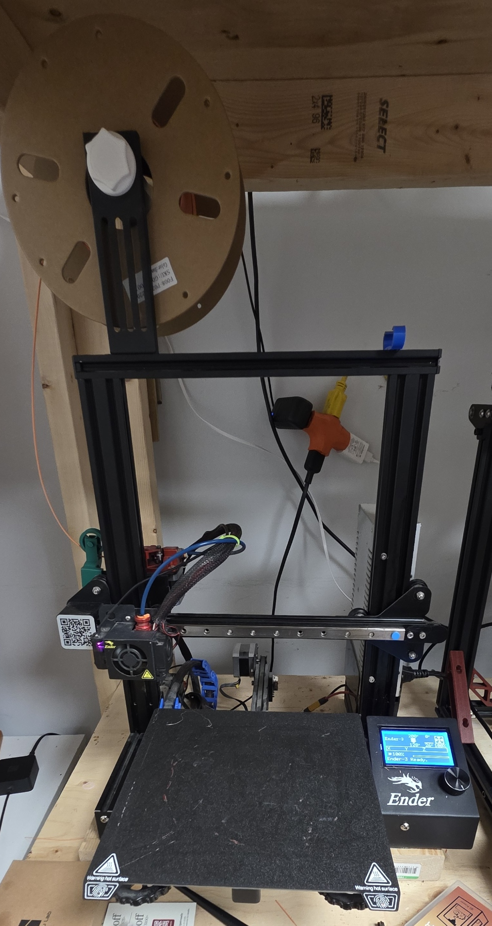 A black 3D printer on a wooden workbench with various accessories, cables, and a spool of filament in the background.